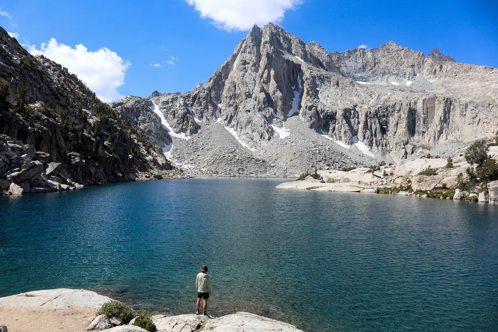 A hiker on a rock at a blue alpine lake under granite peaks.