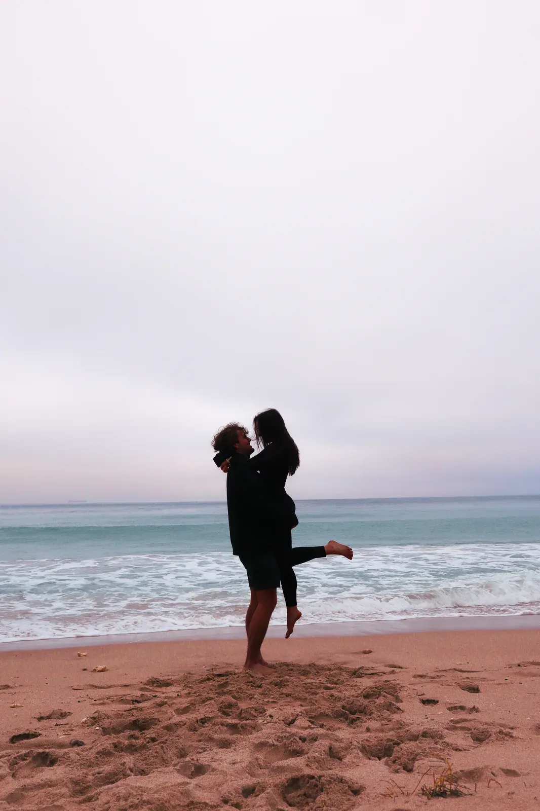 One of them lifting the other in a hug on a beach, waves behind.