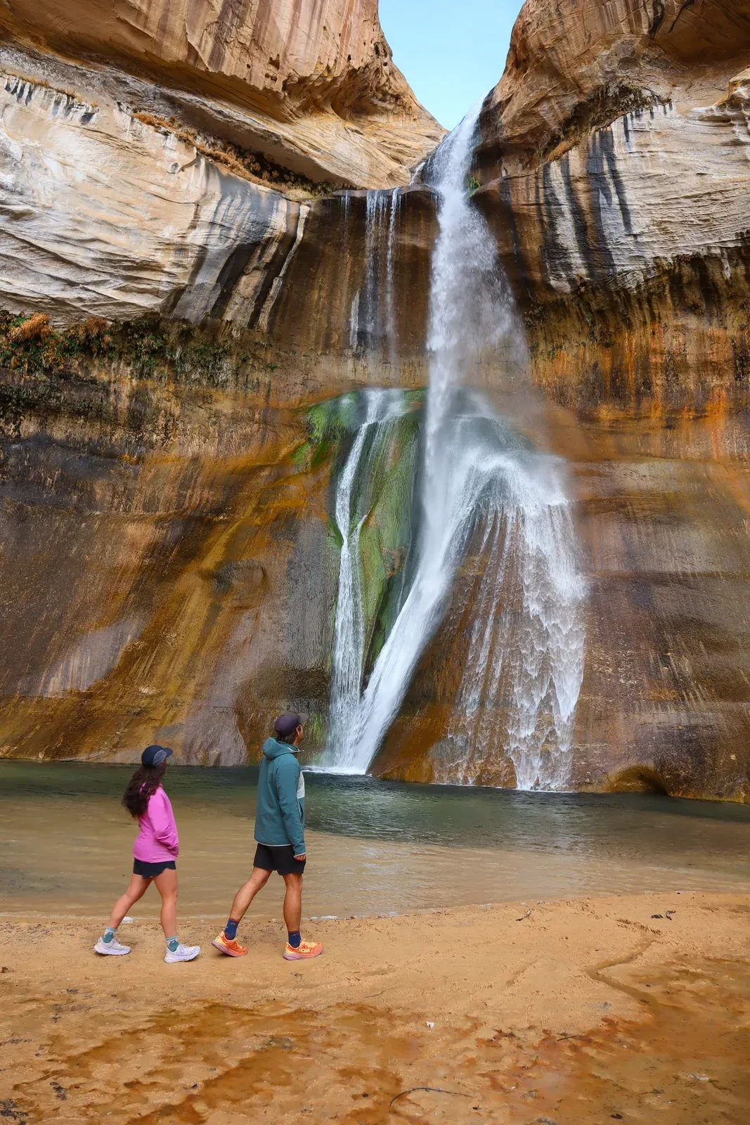Two people wading at the foot of a tall ribbon waterfall on streaked canyon walls.