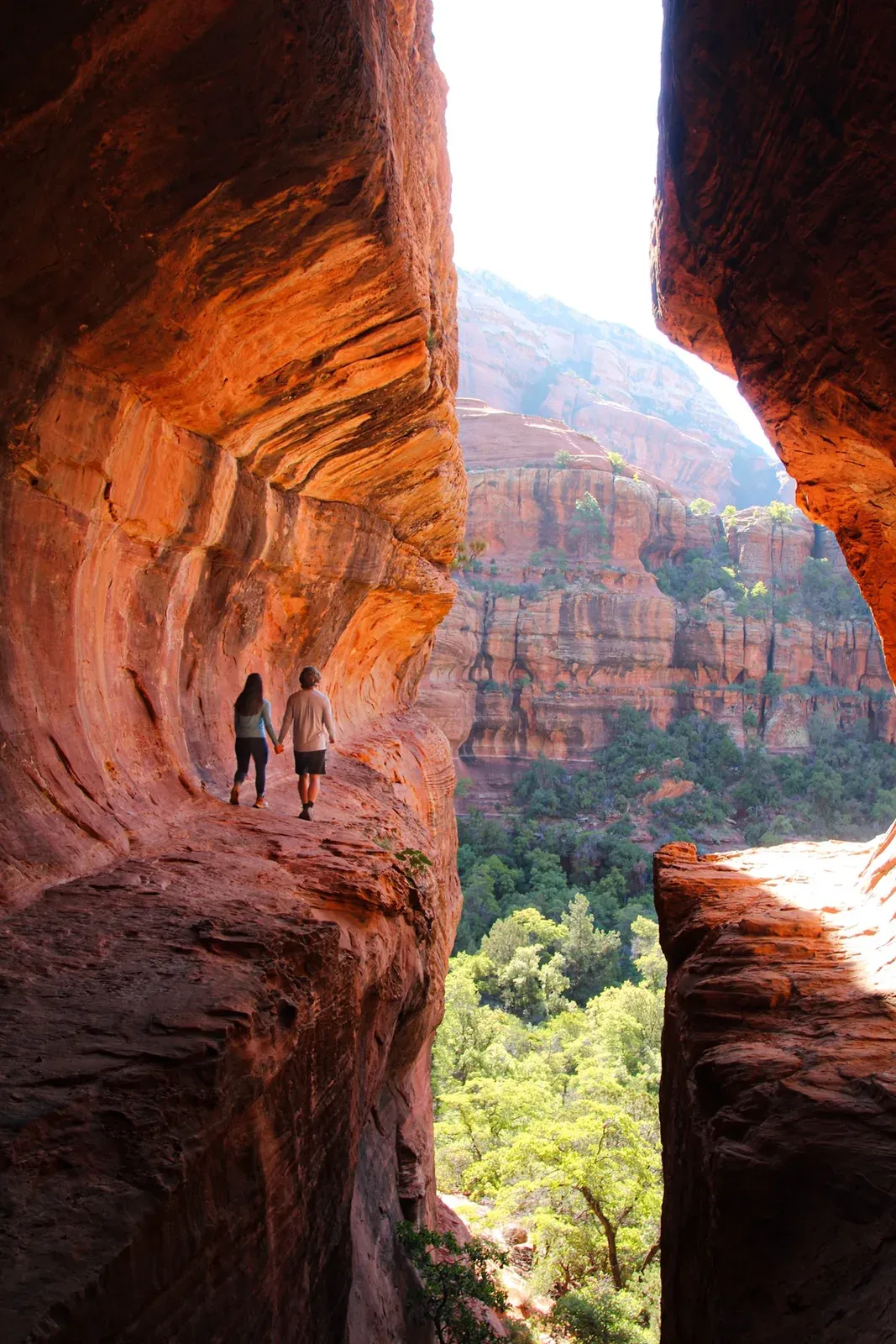 Brendon and Kaylee walking a narrow red-rock ledge inside a cave mouth, a green canyon below.