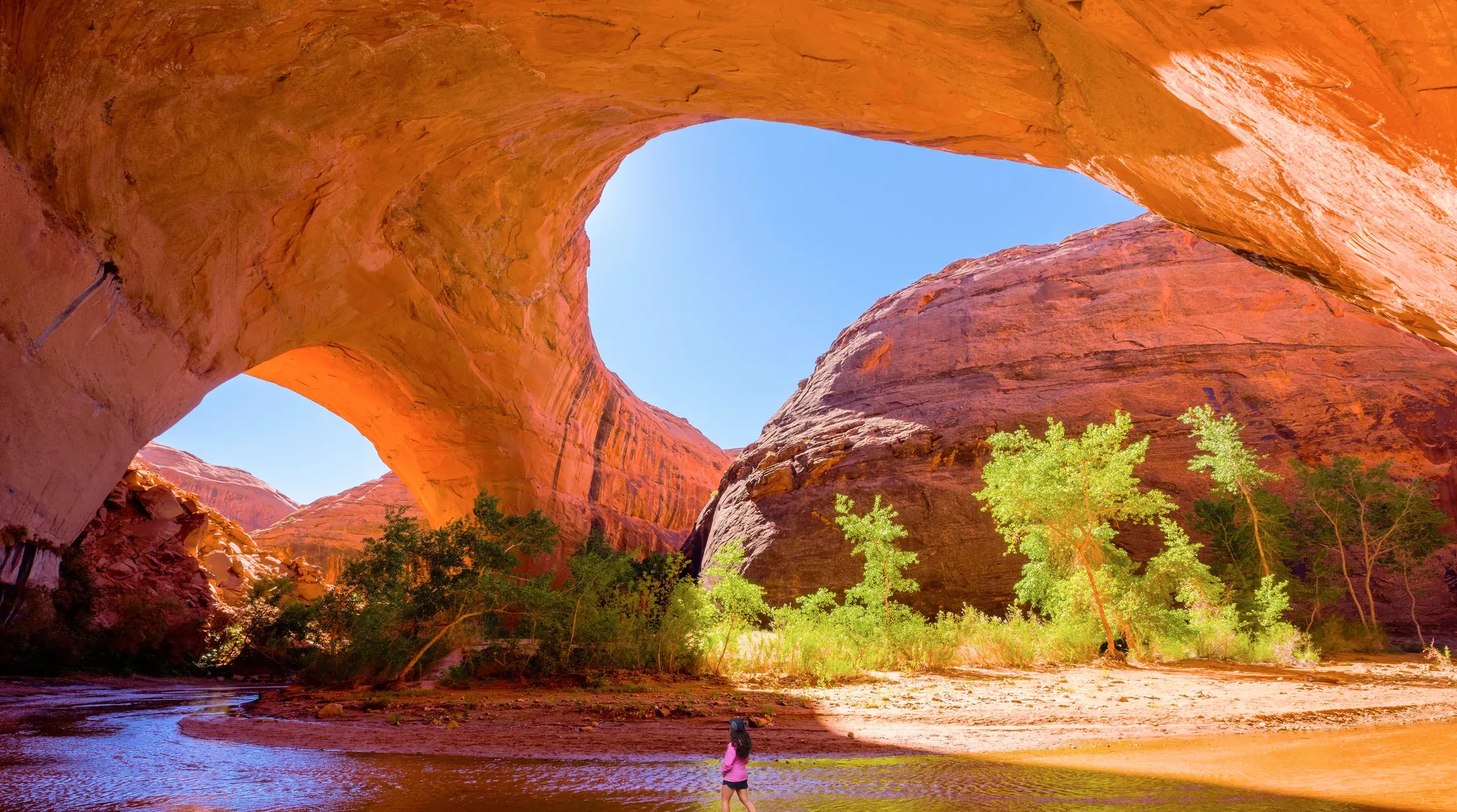 A huge sandstone arch over a creek in Coyote Gulch, green trees below and a small figure in the water.