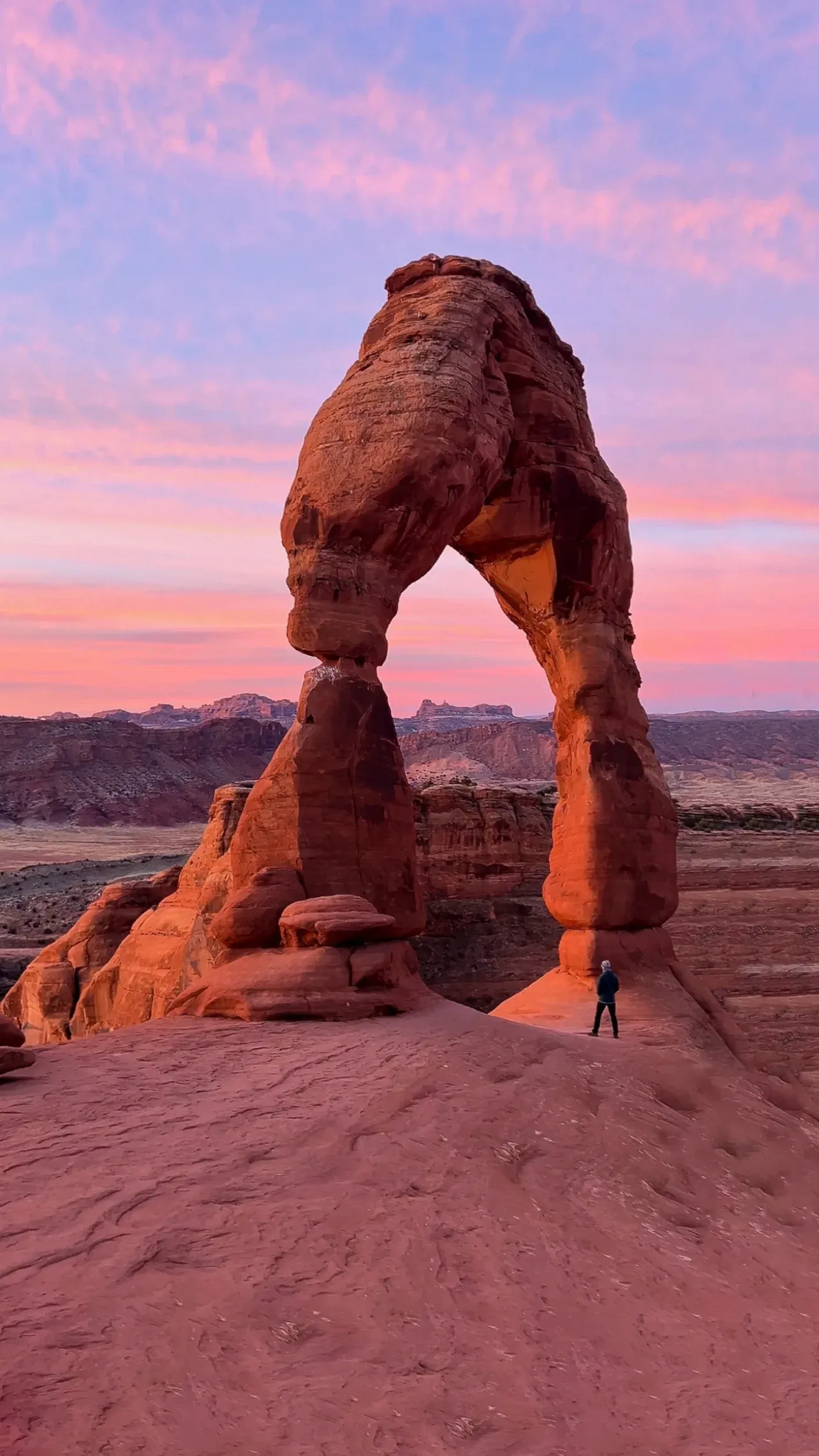 Delicate Arch against a pink and violet sunset sky, a tiny person at its base.