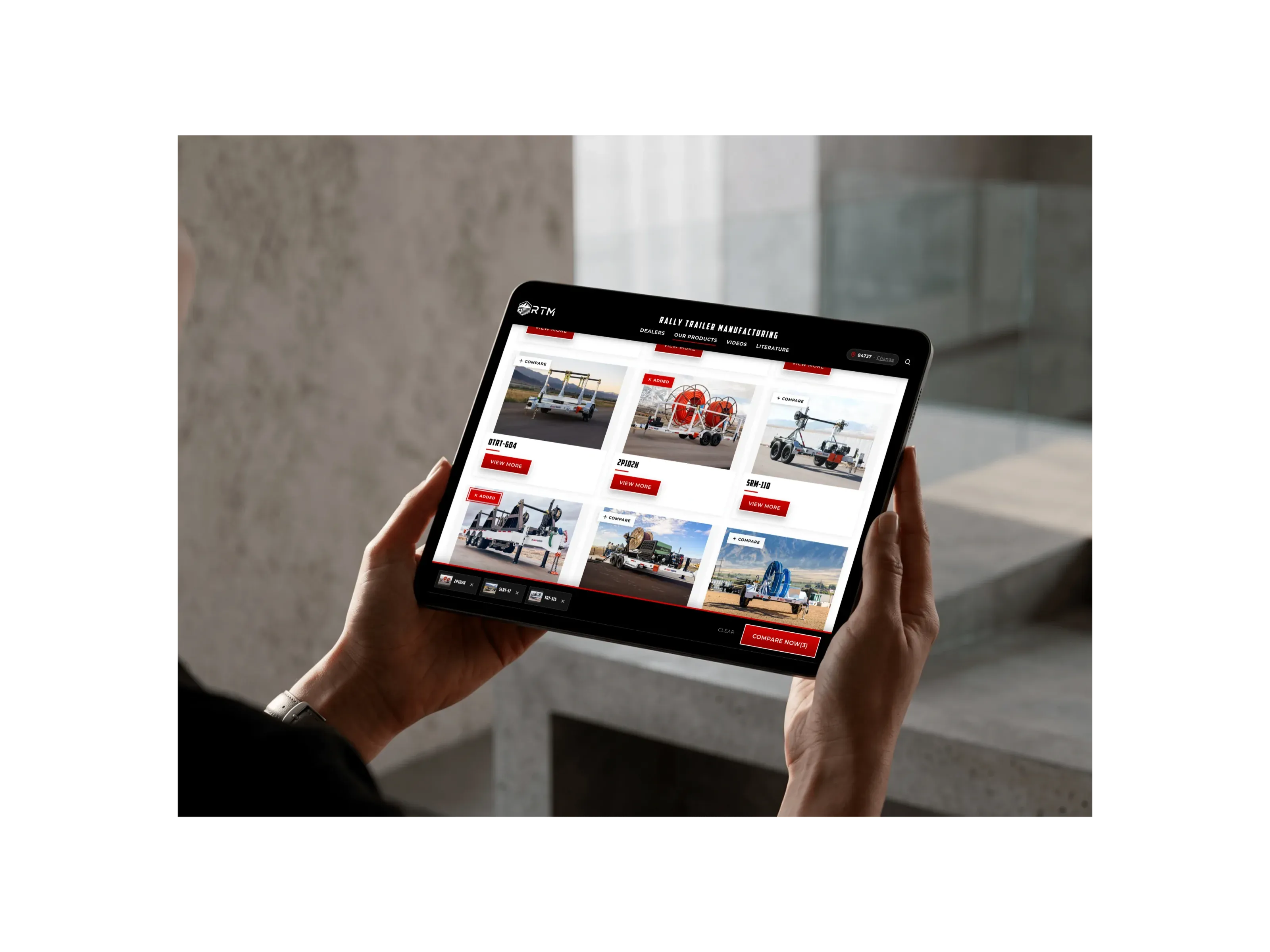 Hands holding an iPad Pro showing the new trailer grid, with compare checkboxes on each trailer and the compare bar along the bottom.