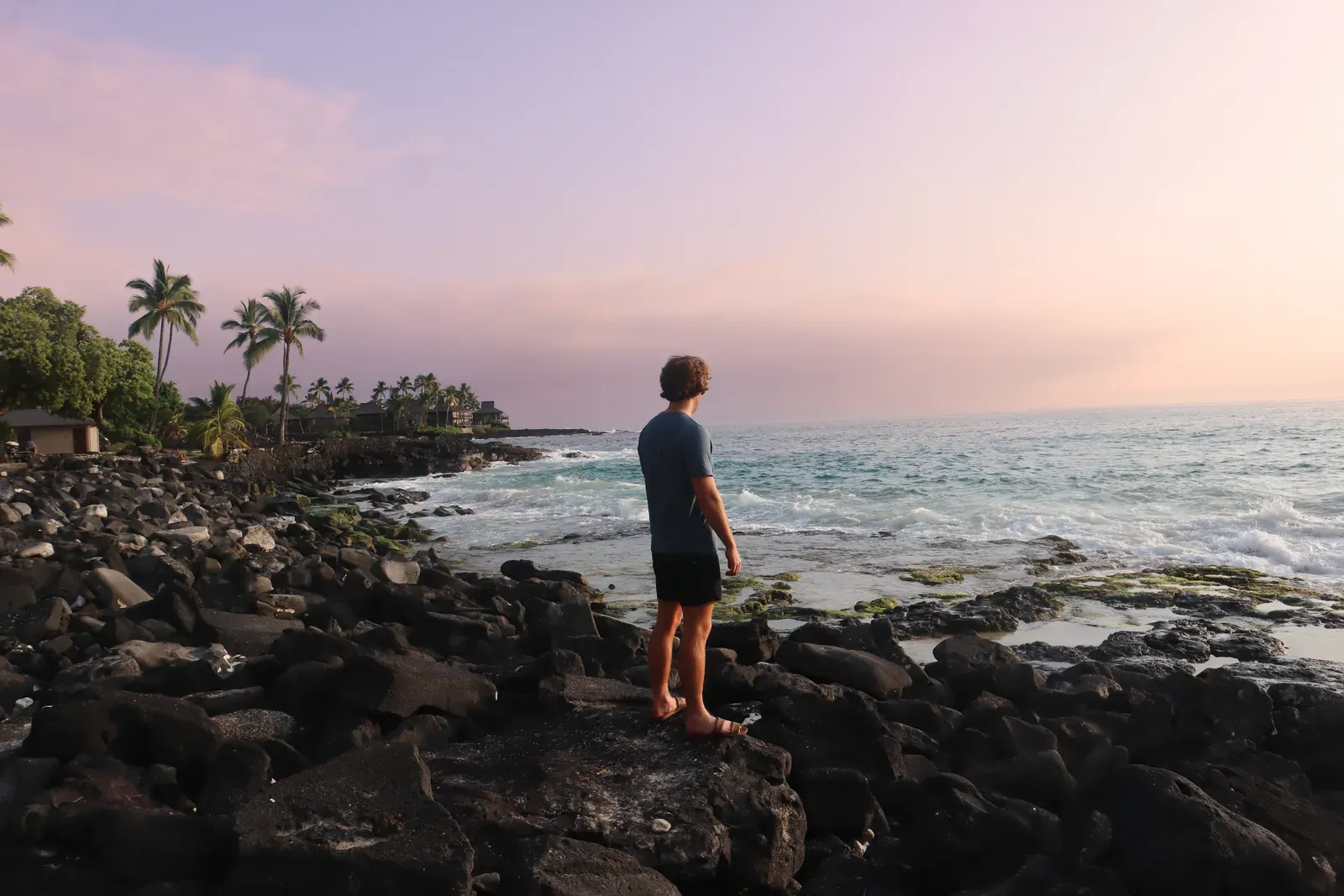 A man standing on black lava rocks by the ocean at dusk, palms behind.
