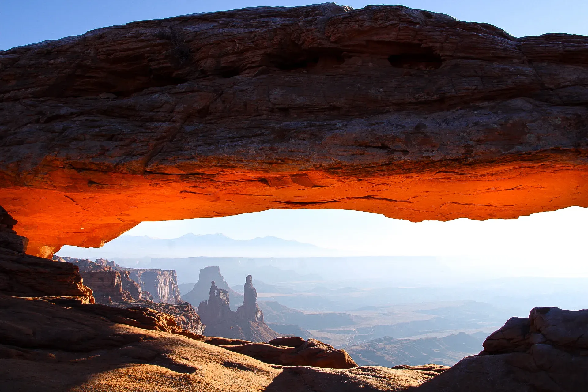 Mesa Arch glowing orange at sunrise, framing towers and canyon below.