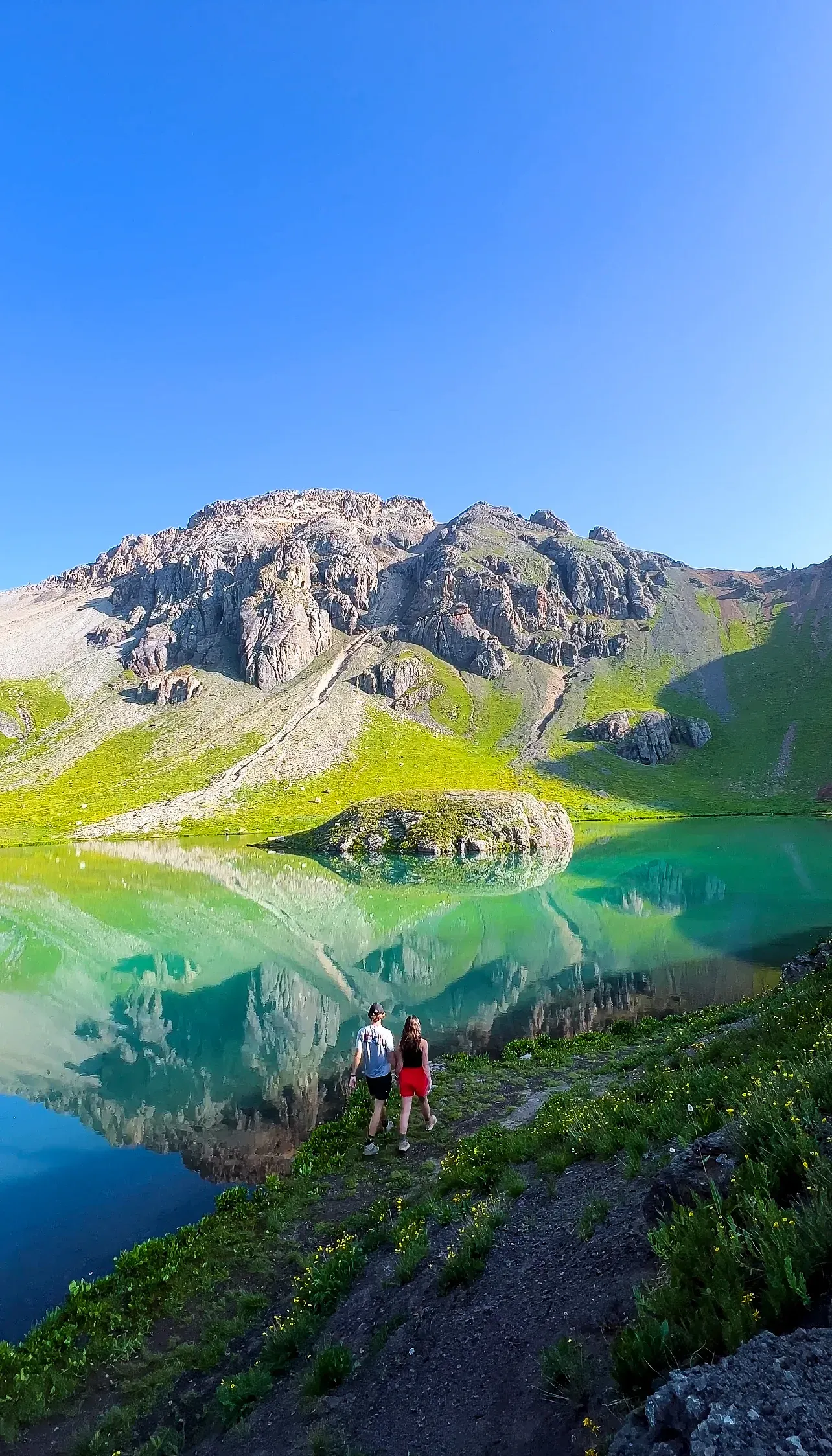 Brendon and Kaylee on a slope above a lake that mirrors a green peak.