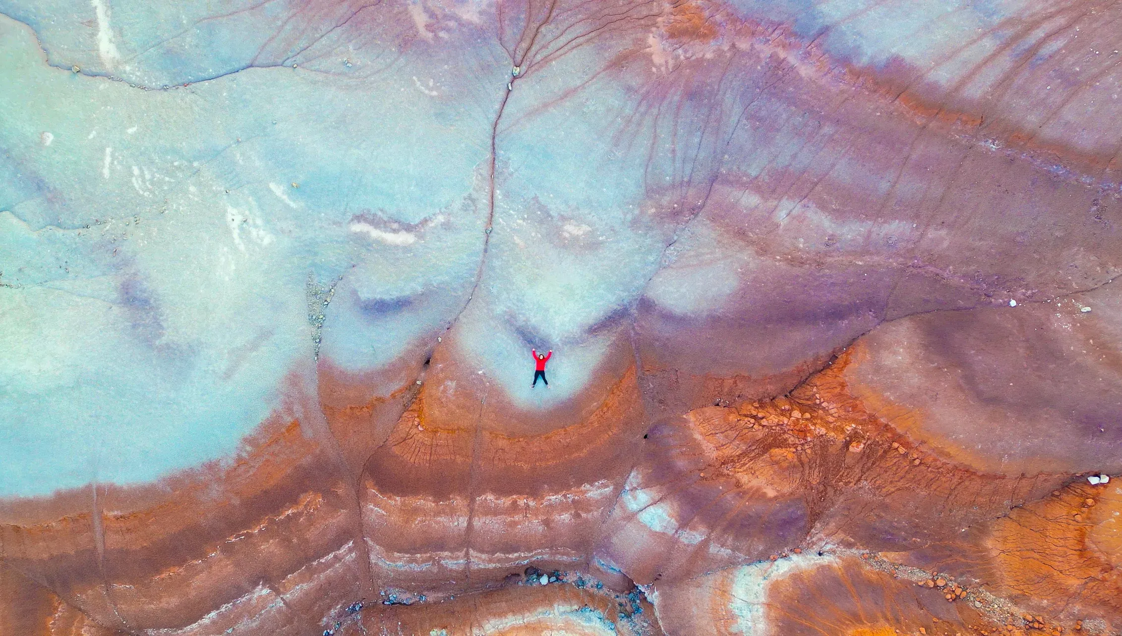 Drone view of the Bentonite Hills: swirling purple, blue and orange clay with a tiny person lying in it.
