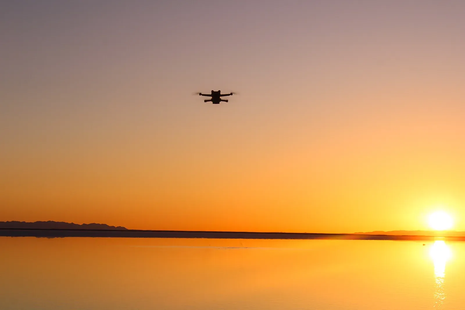 A drone silhouetted over a flat lake at sunset.