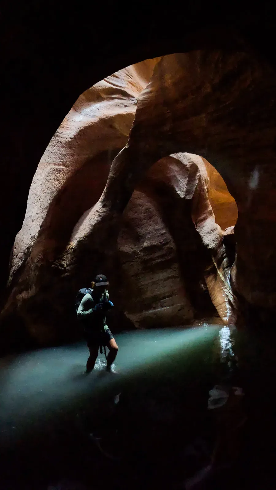 A hiker lit by a shaft of light in a dark curved slot canyon over green water.