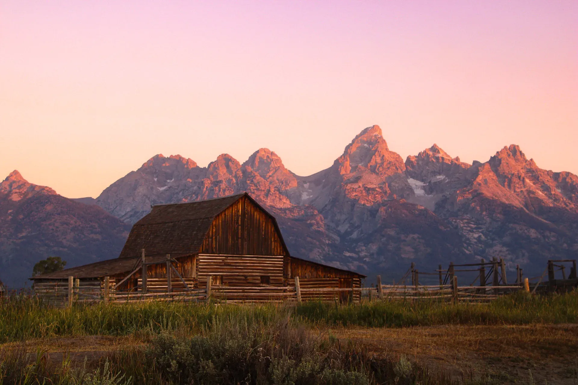 A weathered barn under pink-lit Teton peaks at sunrise.