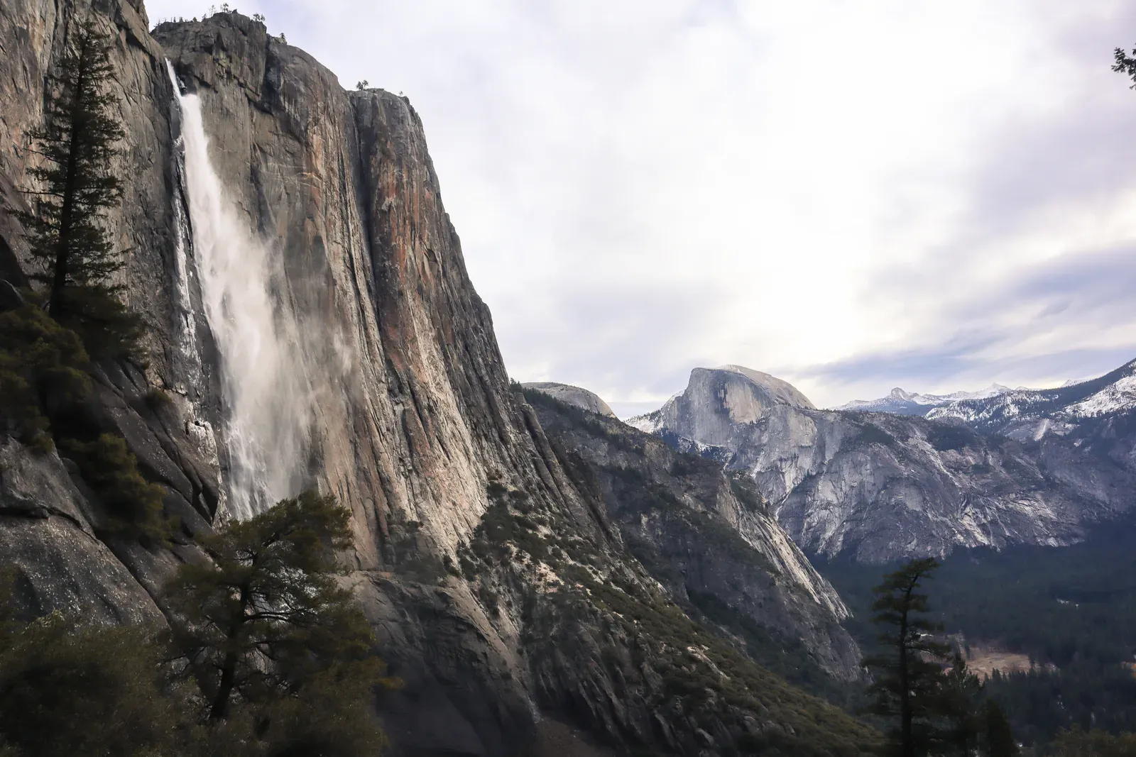 A tall waterfall off a granite wall with Half Dome behind.